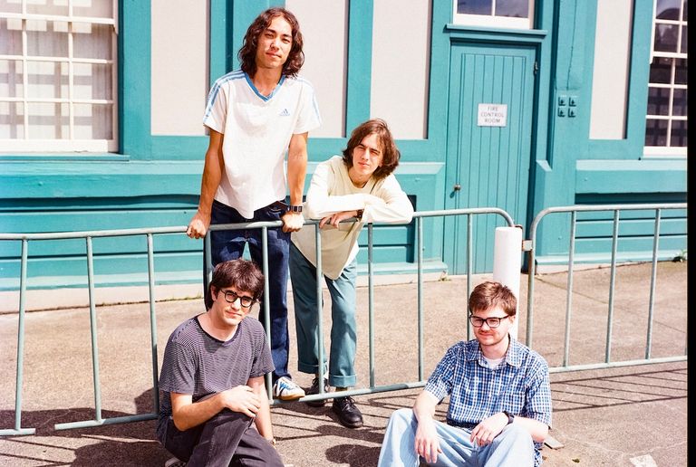 Australian psych-rock band The Lazy Eyes posing together by a metal fence outdoors in front of a teal building for a promo portrait photo.